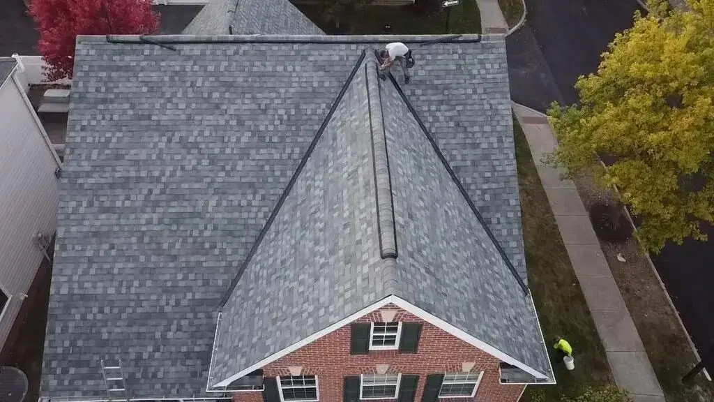 Roofer finishing the ridge cap on a new gray shingle roof over a brick colonial, seen from above
